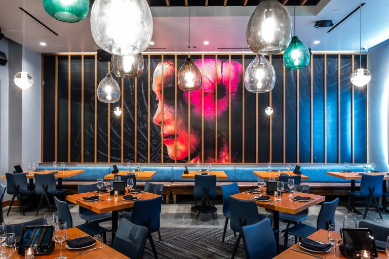 Interior of the Kona Grill Riverton location–a modern design with glass chandeliers, a wall of a mural of a woman's face wearing a large pink flower in her hair.