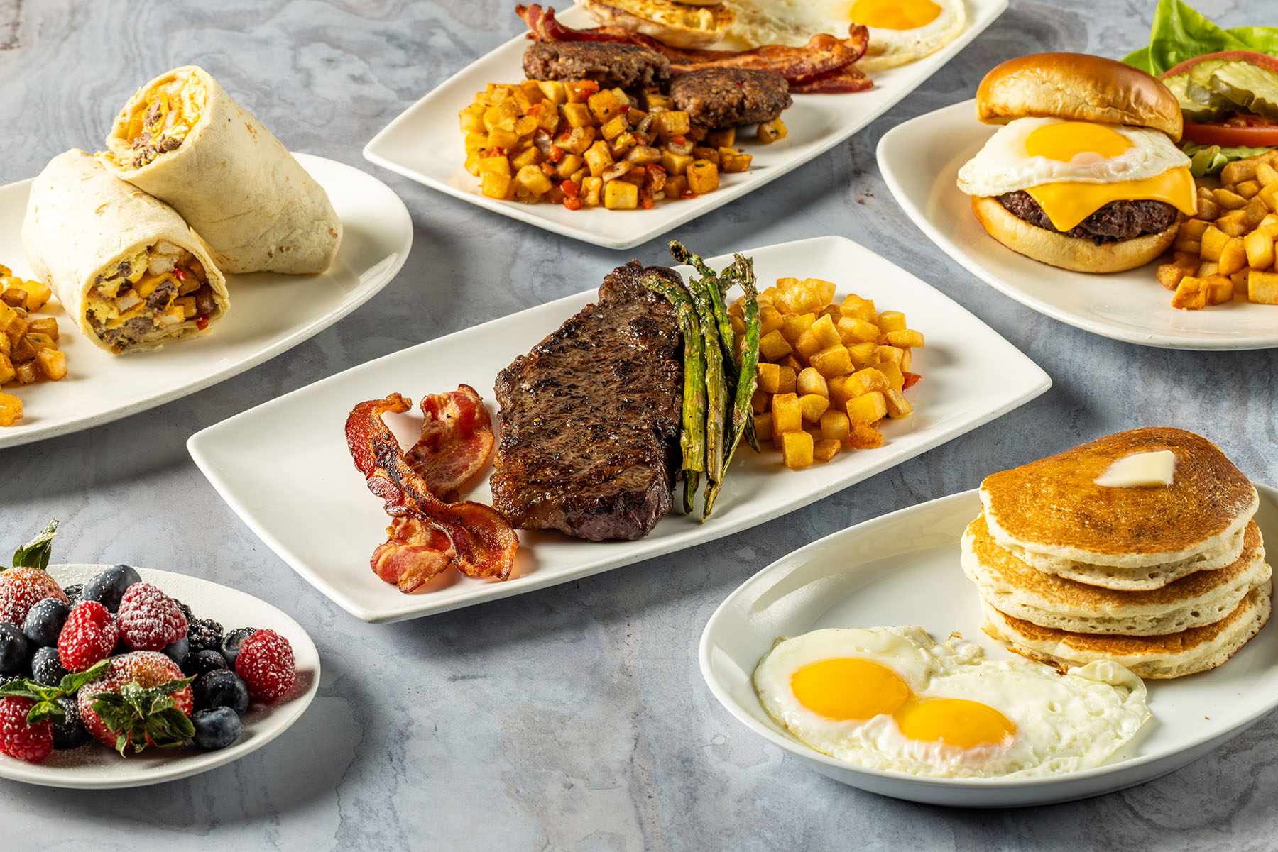 Plates full of Kona Grill brunch dishes including pancakes, fried eggs and bacon, steak and asparagus, breakfast burrito, mixed fresh berries with powdered sugar.