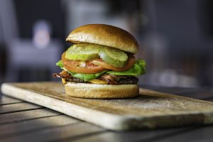 Kona Grill lunch burger with beef, cheese, bacon, lettuce, tomatoes, and pickles on a wooden cutting board