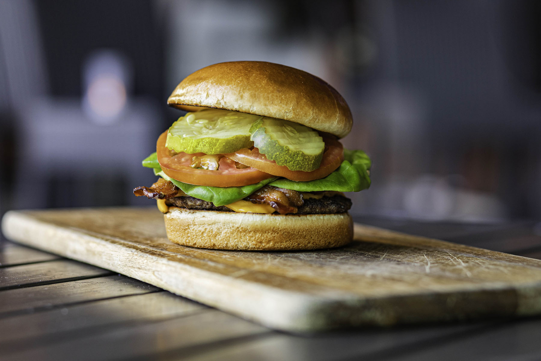 Kona Grill lunch burger with beef, cheese, bacon, lettuce, tomatoes, and pickles on a wooden cutting board